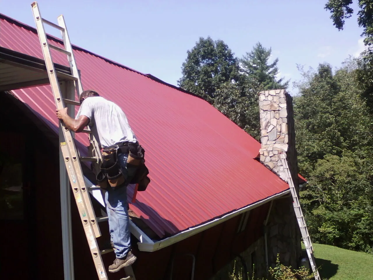 Roofer on a ladder beside a red metal roof and stone chimney — Soffit & Fascia Repair by Roofing Brothers in Jessup